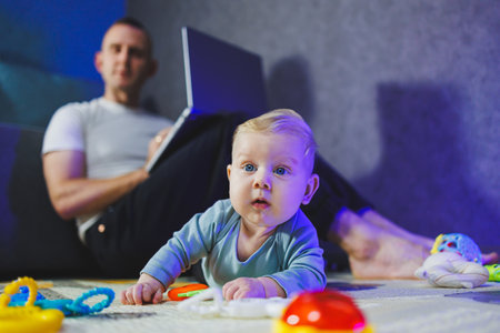 A worried dad is working on a laptop at home while his little son is playing next to him. A baby is playing on the floor.の写真素材