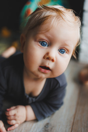 A blue-eyed baby lies on the floor and looks into the camera. A baby with white hair in a gray romper is playing at home. Clothes for babies.の写真素材