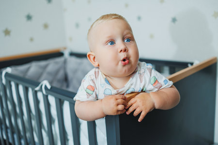 A blue-eyed baby in a t-shirt stands by the railing in his crib. Emotions of the baby. Cot for a...