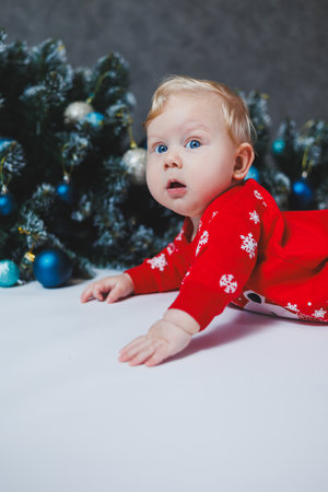 A cute baby in a red Christmas romper is lying near a Christmas tree. Christmas baby. Holiday clothes for childrenの写真素材