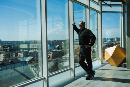 Smiling old mature businessman in stylish glasses and black shirt standing near window in modern...