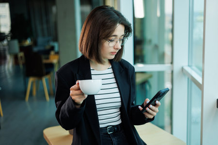 Concentrated business woman wearing glasses while working on a laptop computer while sitting in her spacious office with a cup of coffee on the table.の写真素材