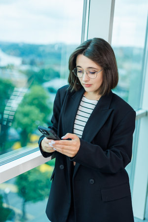 A pretty woman in glasses is standing by the window in a transparent office. A woman rests looking out the window in the officeの写真素材