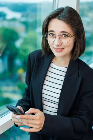 A pretty woman in glasses is standing by the window in a transparent office. A woman rests looking out the window in the officeの写真素材