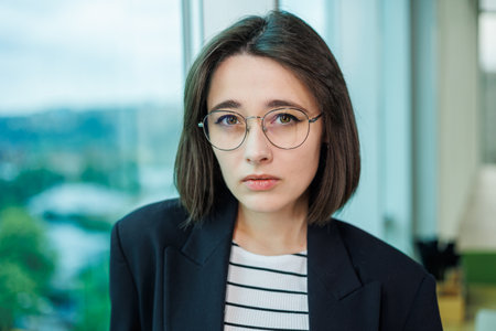 A pretty woman in glasses is standing by the window in a transparent office. A woman rests looking out the window in the officeの写真素材