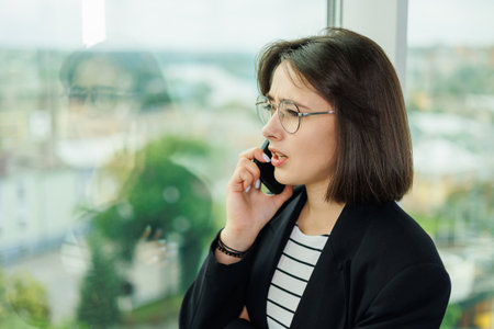 A pretty woman in glasses is standing by the window in a transparent office. A woman rests looking out the window in the officeの写真素材