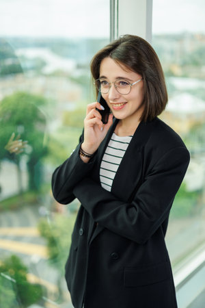 A pretty woman in glasses is standing by the window in a transparent office. A woman rests looking out the window in the officeの写真素材