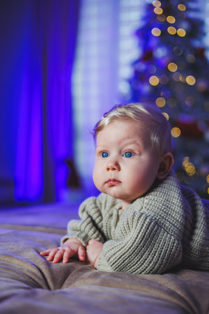 A baby in a beige knitted sweater on the background of a Christmas tree. Baby's first Christmas. A small child on the background of Christmas decorations.の写真素材