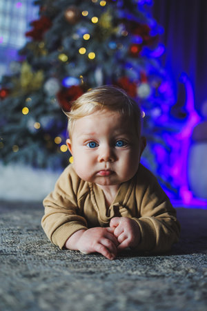 A blue-eyed baby lies on the floor against the background of a Christmas tree. A child at home in a...