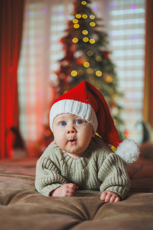 A baby boy in a Santa Claus hat on the background of a Christmas tree. A baby in a knitted sweater.