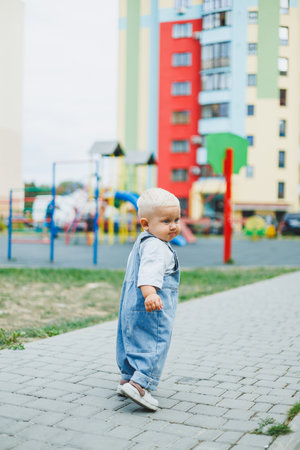 A 1-year-old baby boy in denim overalls is walking on the street. Children's oversized overalls. Stylish little boy on the playground.の写真素材