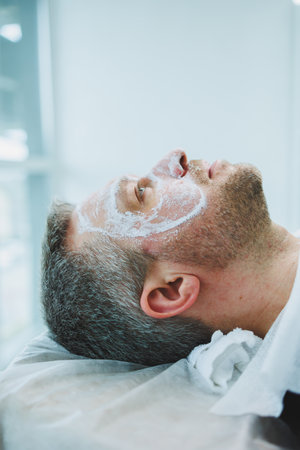 A man with a mask in a beauty salon. Men's face care concept. A beautician puts a mask on a client's face in a spa center. Male cosmetology.の写真素材