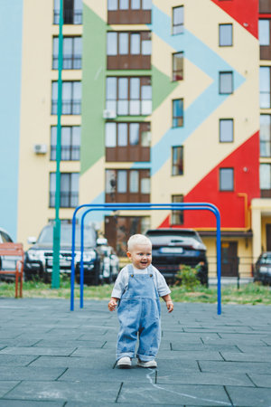 A toddler boy in denim overalls runs and plays on the playground. Active time of the child outside. A child runs on the street in the summer.の写真素材
