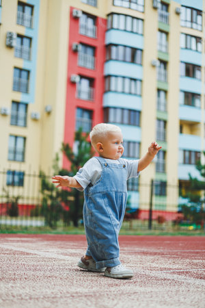 A toddler boy in denim overalls runs and plays on the playground. Active time of the child outside. A child runs on the street in the summer.の写真素材
