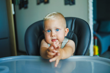 A little baby boy is sitting in a high chair. An emotional child sits in a chair and eats. Feeding a child.の写真素材