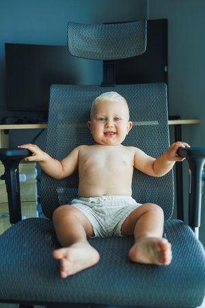 Toddler boy in shorts sits in a comfortable chair. The baby laughs and sits on a computer chair.の写真素材
