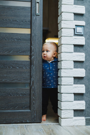 Blond toddler boy at home peeks into the room through a crack in the door. Boy 1 year old opens the door to the room. Children at home.の写真素材