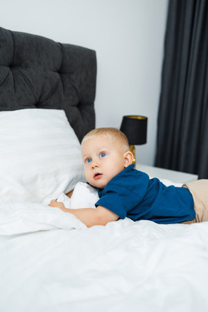 A blond one-year-old boy in a blue T-shirt lies on a bed with white sheets. The baby plays while sitting in bed. A blue-eyed little boy.の写真素材