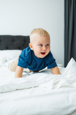 A blond one-year-old boy in a blue T-shirt lies on a bed with white sheets.