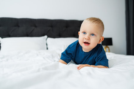 A blond one-year-old boy in a blue T-shirt lies on a bed with white sheets. The baby plays while sitting in bed. A blue-eyed little boy.の写真素材