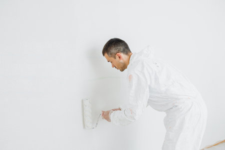 Man doing house renovation. Industrial theme. Painting walls. Builder in white overalls paints wall with white paint roller.の写真素材