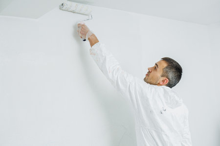 Man doing house renovation. Industrial theme. Painting walls. Builder in white overalls paints wall with white paint roller.の写真素材