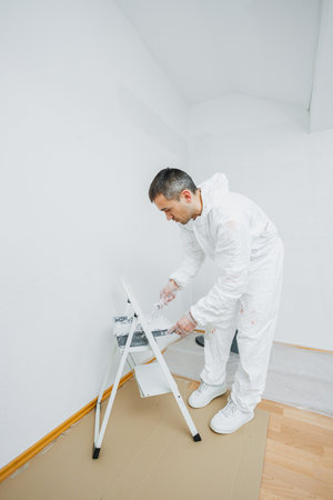Renovation of the premises. A young man with dark hair paints the wall white in the apartment. He holds a roller in his hands.の写真素材