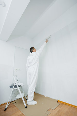 Renovation of the premises. A young man with dark hair paints the wall white in the apartment. He holds a roller in his hands. The concept of cheap repairs.の写真素材