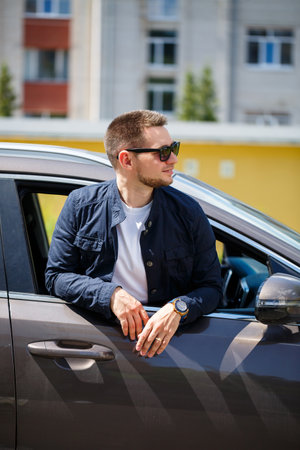 A man, stylishly adorned with sunglasses, leans out of the open window of a car, enjoying the breeze...