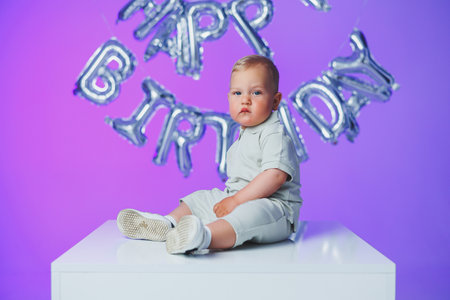 An adorable, cute baby is joyfully sitting on a pedestal, surrounded by colorful Happy Birthday balloonsの写真素材