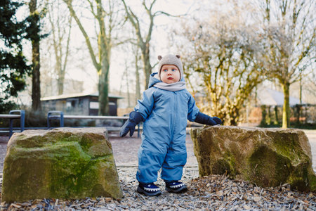 A joyful and curious toddler, dressed warmly in a colorful winter outfit, happily explores a park...