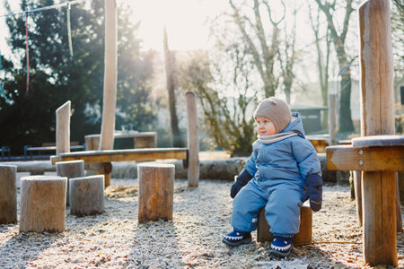 A joyful and cheerful toddler happily sits on a wooden stump in an outdoor play area, warmly dressed for winterの写真素材