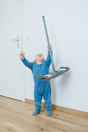 A cute little toddler in charming blue overalls is happily playing with a mop while exploring at...