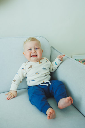 An adorable baby boy is happily smiling while sitting comfortably and cozily on a soft couch with joyの写真素材