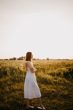 A woman wearing a flowing white dress is relishing a peaceful moment while standing in a sunlit field, enjoying natureの写真素材