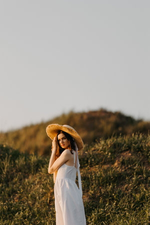 A beautiful woman in an elegant white dress and stylish straw hat stands gracefully in a serene outdoor settingの写真素材