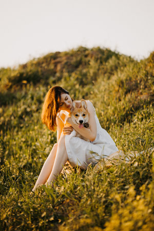 Beautiful girl in a white dress and hat in the field with her dog. Corgi dog on the background of green grass. Girl hugs her corgi dog. Dog walking, friendship. Summer pictureの写真素材