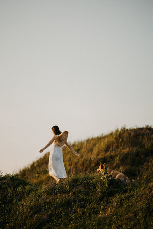 Beautiful girl in a white dress and hat in the field with her dog. Corgi dog on the background of green grass. Girl hugs her corgi dog. Dog walking, friendship. Summer pictureの写真素材