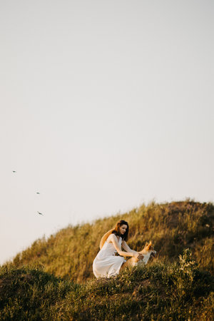 Beautiful girl in a white dress and hat in the field with her dog. Corgi dog on the background of green grass. Girl hugs her corgi dog. Dog walking, friendship. Summer pictureの写真素材