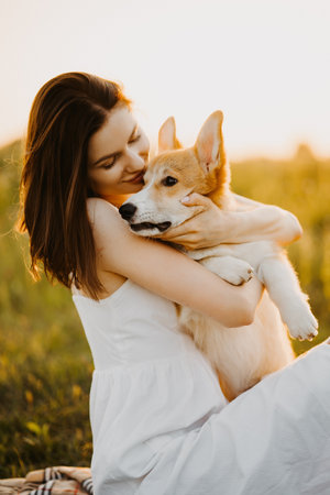 Cute woman in a dress and hat playing with her dog in the field at sunset. Corgi dog on a background of green grass. Girl gently strokes her corgi dog. Dog walking, friendship. Summer pictureの写真素材