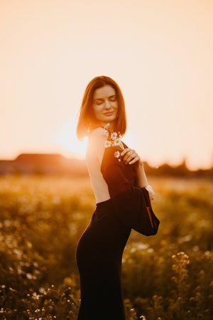 Cute woman in a black dress in a field at sunset. Young woman in nature in a delicate dress basking in the rays of the setting sun.の写真素材