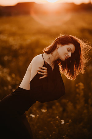 Cute woman in a black dress in a field at sunset. Young woman in nature in a delicate dress basking in the rays of the setting sun.の写真素材