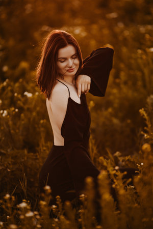 Beautiful woman in a black dress walking in a meadow through the grass at sunset. Woman in a dress in a field at sunset. Woman in nature in a delicate dress basking in the rays of the setting sun.の写真素材