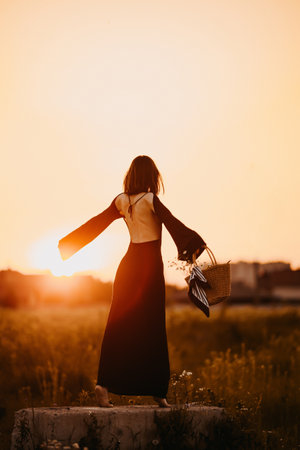 Beautiful woman in a black dress walking in a meadow through the grass at sunset. Woman in a dress in a field at sunset. Woman in nature in a delicate dress basking in the rays of the setting sun.の写真素材