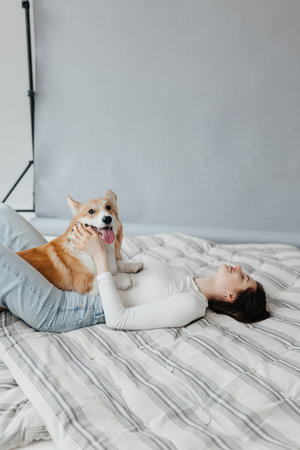 A joyful and warm interaction takes place between a woman and her equally playful corgi on a cozy bedの写真素材
