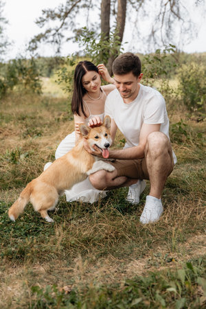 A charming couple enjoys a sunny day outdoors with their playful Corgi in a beautiful natural setting.の写真素材