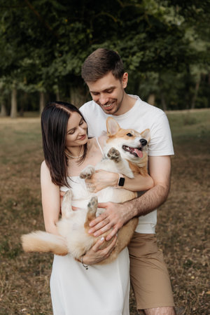 A joyful couple happily holding a playful Corgi in a serene and picturesque outdoor setting surrounded by natureの写真素材