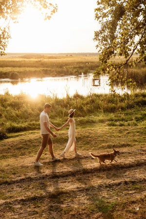 A romantic scene of a couple enjoying a walk with their dog by a serene lake at sunset during golden hourの写真素材