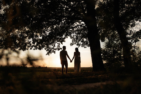 A beautiful silhouette of a couple holding hands against a stunning sunset, surrounded by vibrant treesの写真素材