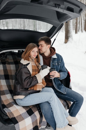 A fashionable couple enjoying each others warmth and savoring drinks together in a snowy landscape from their cozy carの写真素材